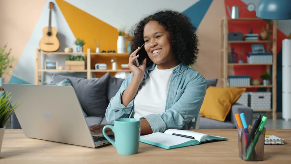 A woman smiling on a phone call at a desk with a laptop and an open notebook