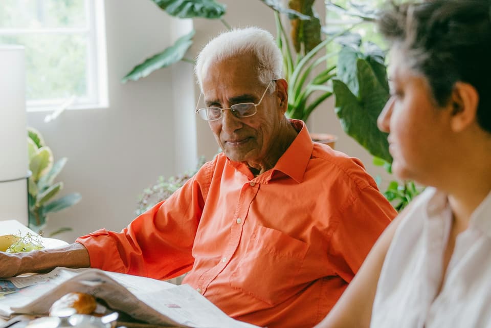 An older man reading a newspaper at a table at home, with a younger woman beside him