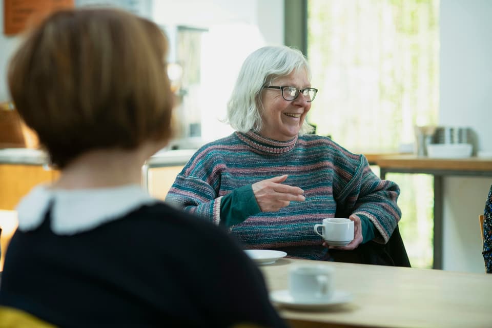 An older woman smiling over a cup of coffee as she talks with a younger woman